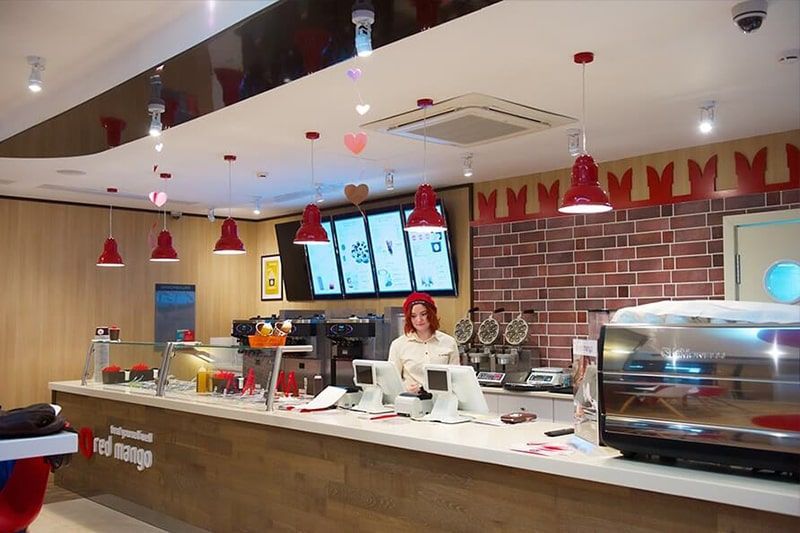 Барная столешница в кафе Red Mango, выполненная из искусственного камня с отделкой ламинатом цвета ясеня. На поверхности расположены витрины с ингредиентами, кассовая зона и кофе-машина.